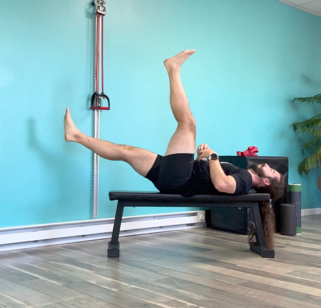 Dr. John laying with his back flat on a bench, right leg up towards the ceiling, left leg lowering down and extending forward. This exercise helps coordinate your core and hamstring movement to heal your hamstring strain fast